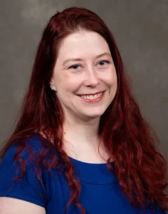 Colleen Baldwin is smiling and has long red hair. Colleen wears a blue shirt.