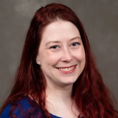 Colleen Baldwin is smiling and has long red hair. Colleen wears a blue shirt.