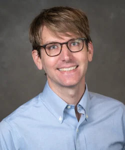 Ryan Powell smiles for his bio photo. He has short light brown hair, light brown glasses, and he wears a light blue button up.