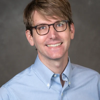 Ryan Powell smiles for his bio photo. He has short light brown hair, light brown glasses, and he wears a light blue button up.