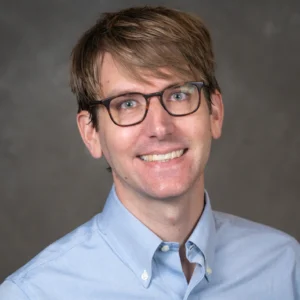 Ryan Powell smiles for his bio photo. He has short light brown hair, light brown glasses, and he wears a light blue button up.
