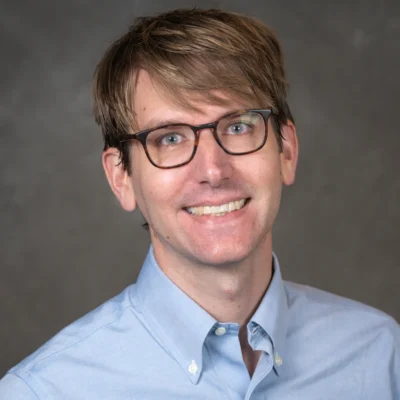Ryan Powell smiles for his bio photo. He has short light brown hair, light brown glasses, and he wears a light blue button up.