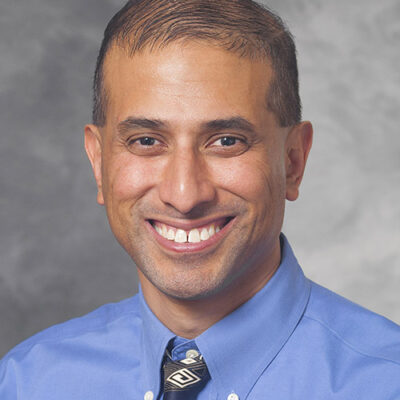 Manish Shah is smiling while wearing light blue. Manish has short, dark hair.