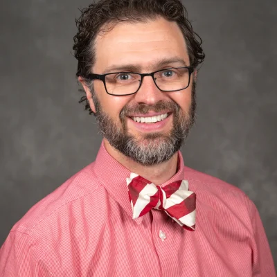 William Buckingham's bio headshot. Will wears a light red/pink shirt and smiles. Will also has a red and white bowtie.