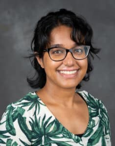 Ruvini Navaratna smiles as she wears a patterned green and white shirt. She has short black hair and black glasses.
