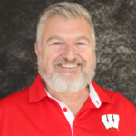 Dave Lyons smiles. He has light grey hair and wears a bright red UW-Madison button up.