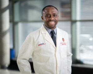 Man wearing a lab coat and tie smiles for a photo.