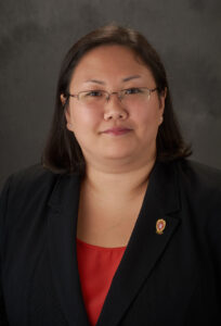 Jenny smiles slightly for her headshot. She has glasses, long dark hair, and wears a professional black blazer.