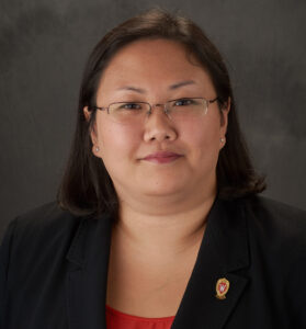 Jenny smiles slightly for her headshot. She has glasses, long dark hair, and wears a professional black blazer.