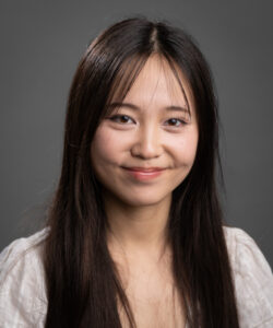 Guiying (Angel) Zhong is a Chinese American woman in their 20s. They have long dark hair and wear a white cardigan.