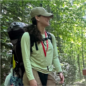 A woman in a baseball cap stands along a hiking trail in the woods.