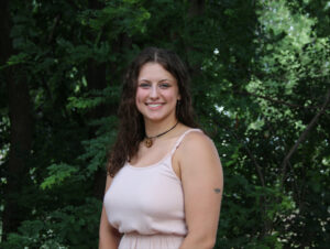 Brunette woman stands in front of foliage.