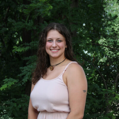 Brunette woman stands in front of foliage.