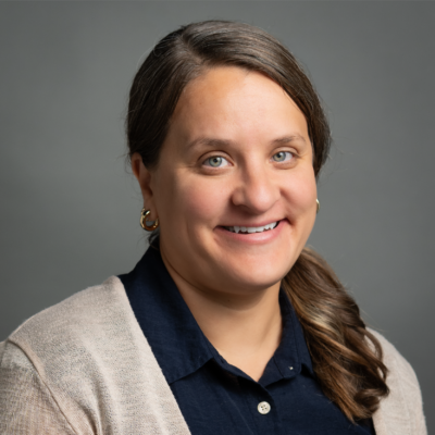 Smiling woman with brown hair in a ponytail, wearing a dark shirt and light cardigan, against a gray background.